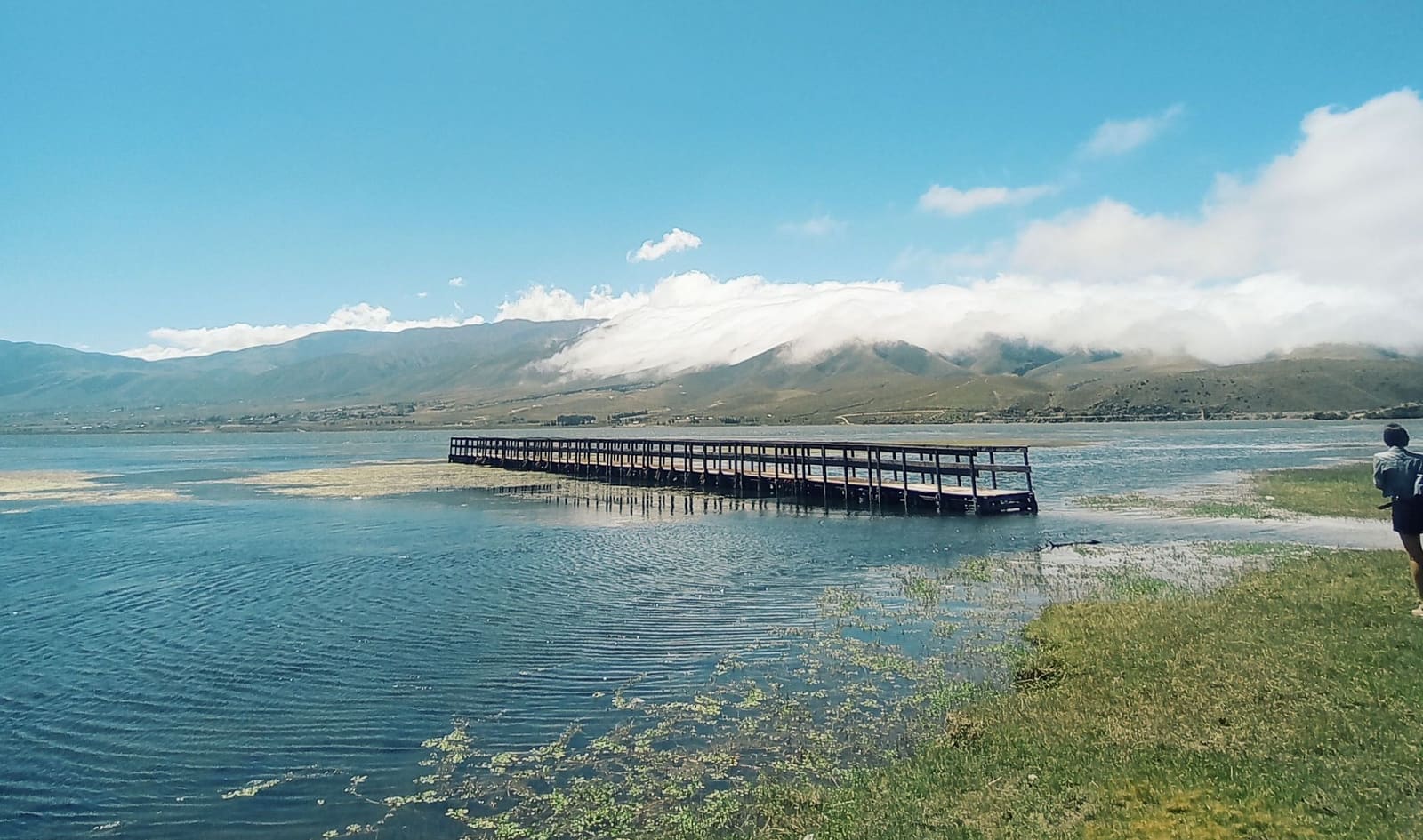 El muelle del dique La Angostura sobre el agua, con los cerros del valle y un mar de nubes al fondo